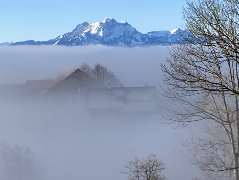 Bauernhof im Nebel vor schneebedecktem Pilatus und blauem Himmel